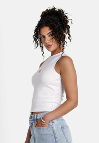 Top de tirantes blanco con cuello alto y diseño sin mangas. Presenta un pequeño logo rojo en el pecho y se combina con unos vaqueros azul claro.
