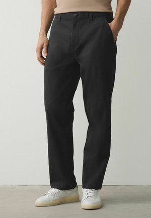 Man wearing black straight-leg trousers with side pockets and white sneakers, standing against a plain light background.