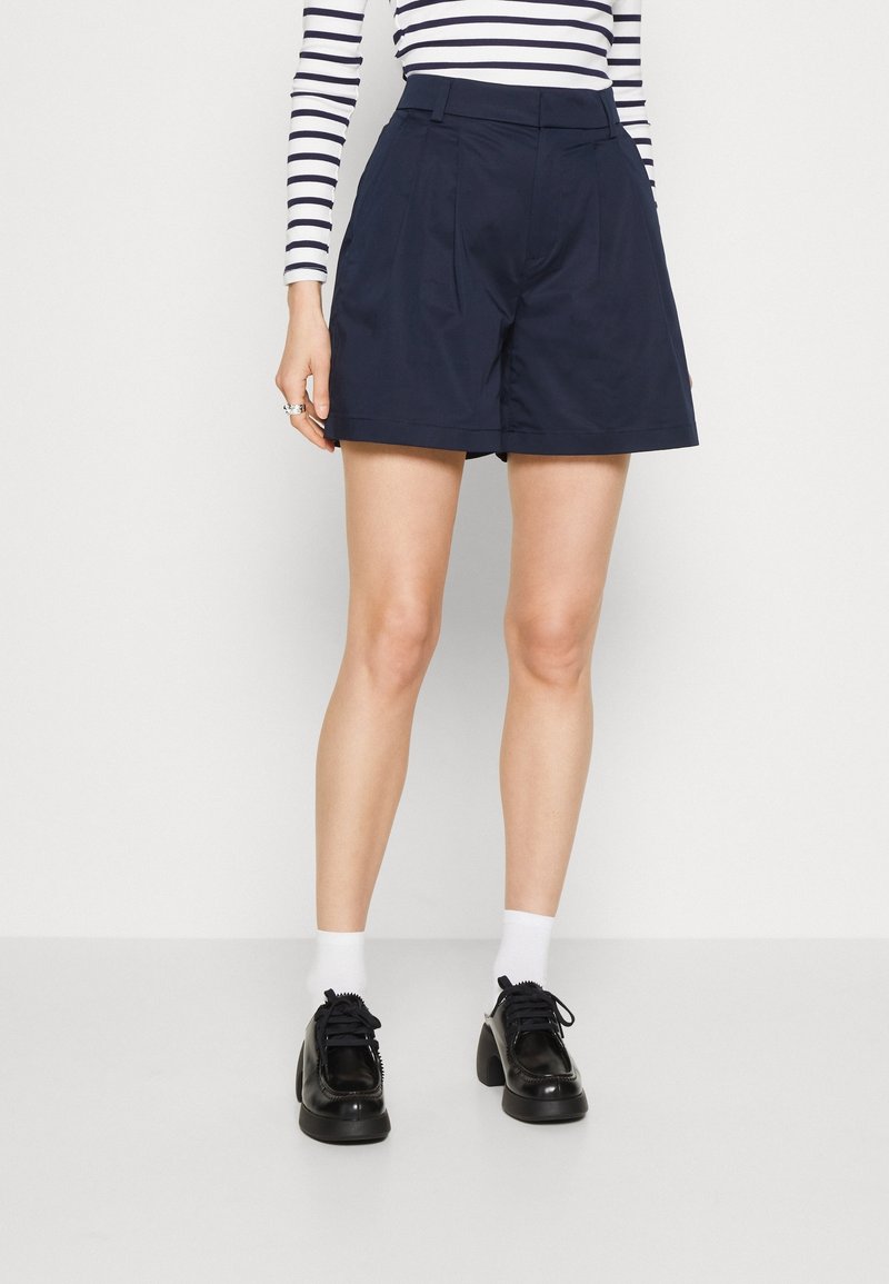 Femme portant un short taille haute tailleur bleu marine, des chaussures noires à talons épais, des chaussettes blanches et un haut blanc à manches longues avec des rayures horizontales noires.