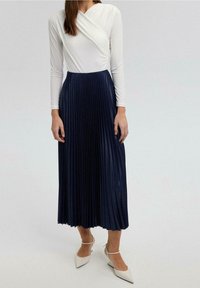 Body a maniche lunghe bianco abbinato a una gonna a pieghe blu navy, con una finitura texturizzata e una silhouette fluida. Tacchi bianchi a punta.