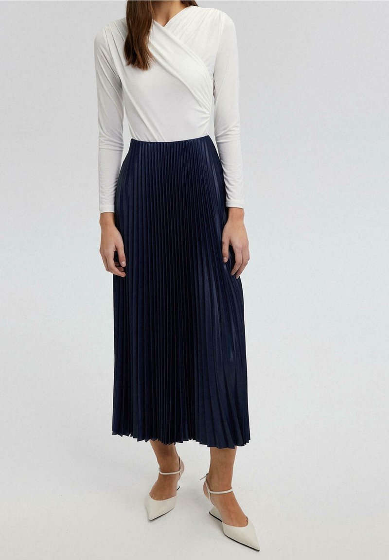 Body a maniche lunghe bianco abbinato a una gonna a pieghe blu navy, con una finitura texturizzata e una silhouette fluida. Tacchi bianchi a punta.