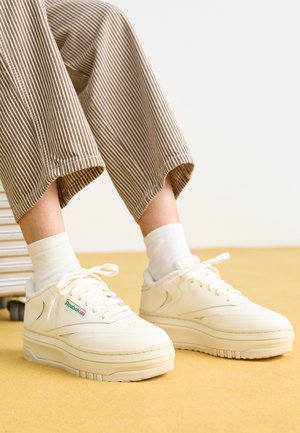 Personne portant des baskets blanches Reebok avec des chaussettes blanches et un pantalon rayé marron, debout sur une surface jaune à côté d'une valise argentée.