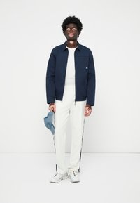 Chaqueta azul marino abotonada con un bolsillo, camiseta blanca y pantalones blancos con franjas laterales en azul marino, completando el look con zapatillas blancas.