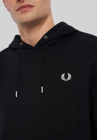 Sudadera negra con bolsillo tipo canguro, cordones con puntas metálicas y un escudo bordado en blanco en el lado izquierdo del pecho. Tejido texturizado.