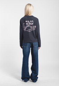 Langärmliges, dunkelgraues Shirt mit einem grafischen Design auf dem Rücken, das Tacos und Getränke in Pink zeigt. Kombiniert mit weit geschnittenen blauen Jeans.