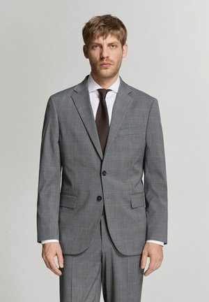 Mann mit hellbraunen Haaren, der einen grauen karierten Anzug, ein weißes Hemd und eine dunkelbraune Krawatte trägt, steht vor einem schlichten hellen Hintergrund.