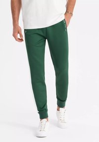 Gröna sportiga joggers i slät tyg, med avsmalnande mudd och sidostripes med varumärkeslogot. Kombineras med vita sneakers.