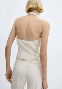 Blonďatá žena v bezrukávovém, bezhřbetém béžovém topu a odpovídajících kalhotách s vysokým pasem, stojící na neutrálním světlém pozadí.