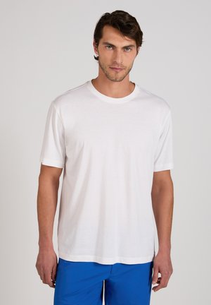 Hombre con cabello oscuro que lleva una camiseta blanca de manga corta lisa y pantalones azules, de pie frente a un fondo gris claro.