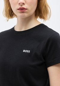 Νεαρό άτομο με κοντά ξανθά μαλλιά, φορώντας μαύρο μπλουζάκι με λευκό κείμενο "BOSS" στο στήθος, μπροστά σε ένα απλό φόντο.