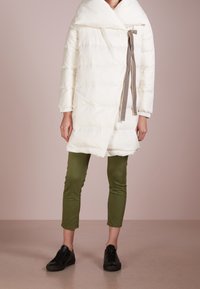 Vit puffjacka med överdimensionerad krage, lätt vaddering och en brun mönstrad snörning. Kombinerad med gröna byxor och svarta sneakers.