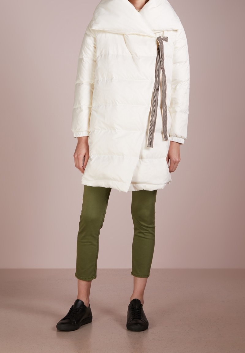 Vit puffjacka med överdimensionerad krage, lätt vaddering och en brun mönstrad snörning. Kombinerad med gröna byxor och svarta sneakers.