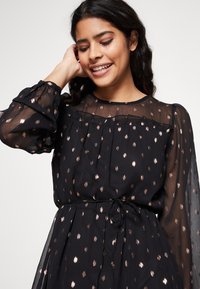 Robe noire avec une superposition transparente, ornée de points en rose doré métallique, manches longues, encolure volantée et taille nouée pour un look ajusté.