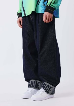 Personne portant un jean sombre ample retroussé pour montrer des revers intérieurs gris avec texte, associé à des baskets blanches et un haut coloré à manches longues.