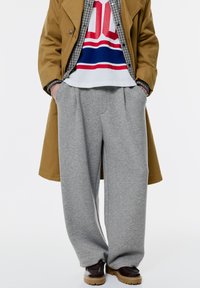 Sive trenirke z ohlapnim krojem in mehkim teksturiranjem, v kombinaciji z rjavim plaščem, barvno grafično majico in temnimi čevlji.