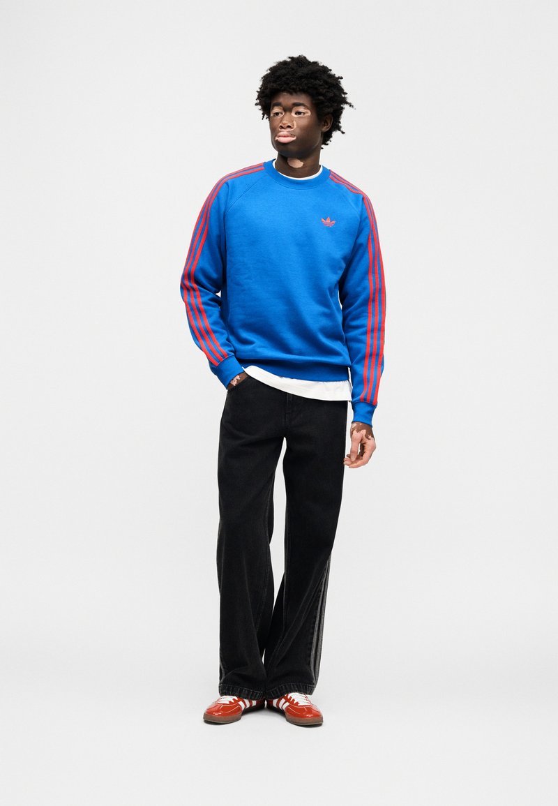 Sweat-shirt bleu avec des rayures rouges sur les manches, jean noir à jambes larges et baskets rouges et blanches. Présente un petit logo sur la poitrine.