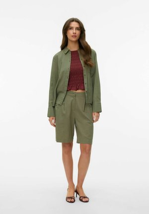 Vrouw staand, gekleed in een olijfgroen overhemd met knopen en knielange shorts met een gestructureerde bordeauxrode top en zwarte sandalen met hak.