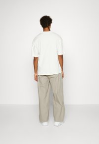 Uomo di spalle che indossa una t-shirt bianca a maniche corte, pantaloni beige a gamba larga e sneakers bianche su uno sfondo chiaro e neutro.
