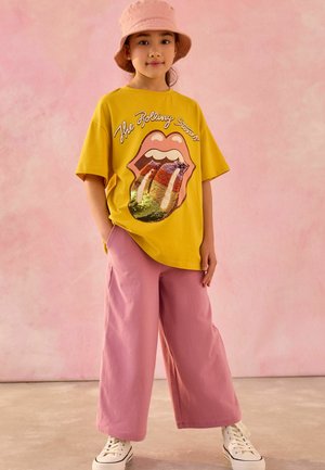 T-shirt en coton jaune avec un logo Rolling Stones brillant, associé à un large pantalon rose et des baskets blanches. Arrière-plan : dégradé rose doux.