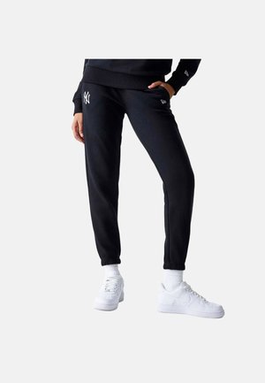 Personne portant un pantalon de survêtement noir avec le logo des New York Yankees, des baskets blanches et des chaussettes blanches, debout avec une main dans la poche.