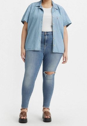 Femme portant une chemise bleu clair à manches courtes boutonnée sur un haut blanc, un jean skinny bleu déchiré et des sandales compensées marron, debout sur fond blanc.