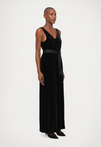 Vestido maxi de terciopelo negro con escote en V y diseño sin mangas, que cuenta con un cinturón a juego en la cintura y un acabado fluido y texturizado.