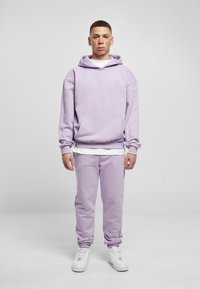 Młody mężczyzna z piegami, ubrany w lawendowy hoodie i dresy, w białych trampkach, stoi na tle jasnoszarej ściany.