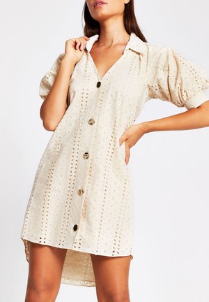 Kobieta w kremowej sukience z ażurowej tkaniny z bufkami, kołnierzykiem i dużymi guzikami z przodu, pozująca z jedną ręką na biodrze.