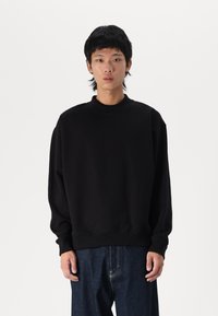 Schwarzes oversized Sweatshirt mit hohem Kragen, gerippten Bündchen und glatter Baumwolltextur, kombiniert mit lockeren dunklen Denim-Hosen.