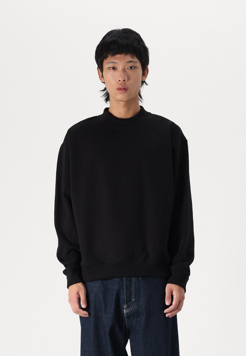 Sort oversized sweatshirt med høj hals, ribbede manchetter og en glat bomuldsstruktur, parret med løse mørke denimbukser.