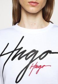 Felpa bianca con un prominente logo cursivo nero "Hugo" e una firma rossa più piccola sottostante. Tessuto morbido e vestibilità classica.