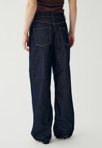 Sötétkék farmer széles lábú farmernadrág magas derekú, hátsó zsebekkel és kontrasztos sárga varrással. Nincs látható fém részlet.