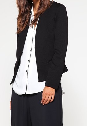 Kvinde iført en tekstureret sort blazer over en hvid skjorte med sorte kanter og løse sorte bukser, vist fra skuldre til hofter.