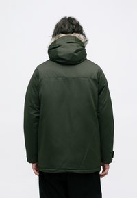 Olivengrøn pufferjakke med hætte og kunstpelskant, der har en glat tekstur og en afslappet pasform, der slutter ved hoften.