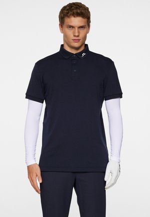 Marineblå polo skjorte lavet af åndbart stof, med knaplukning med tre knapper, korte ærmer og et logo på kraven. Lange hvide ærmer er synlige.