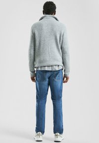 Szary sweter z dzianiny z wysokim kołnierzem, noszony na koszuli w kratę, w połączeniu z niebieskimi dżinsami i białymi trampkami. Widoczna faktura tkaniny.