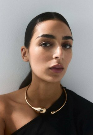 Mujer con el cabello oscuro recogido, vistiendo un top negro de un solo hombro y un collar choker abierto dorado y escultórico, sobre un fondo claro y liso.