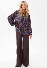 Blonde woman wearing a loose zebra-striped blouse with balloon sleeves, wide brown leather pants, and burgundy pointed shoes.