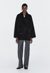 Zwarte dubbelzijdige jas met brede revers en oversized mouwen, gemaakt van een wolmix, gecombineerd met grijze rechttoe rechtaan broek.
