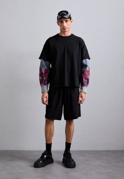 Black oversized t-shirt with flame-patterned sleeves, paired with black shorts and chunky black sneakers. Gray background.