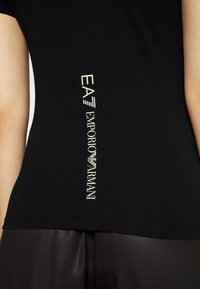 Camiseta negra ajustada de manga corta con estampado vertical en blanco y dorado de "EA7 EMPORIO ARMANI" en el lateral, material de algodón suave.