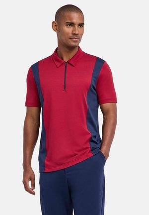 Homme debout les mains dans les poches portant un polo rouge et marine à manches courtes avec une fermeture éclair à l'avant et un pantalon marine.