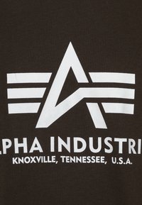 Brunt stof prydes med hvidt trykt tekst og grafik, herunder en abstrakt stjernesymbol og vandrette linjer. Teksten inkluderer "Alpha Industries" samt placeringdetaljer.