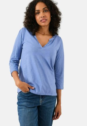 Frau mit lockigem Haar, die ein hellblaues 3/4-Ärmel-Henley-Shirt und blaue Jeans trägt, steht mit einer Hand in ihrer Tasche.