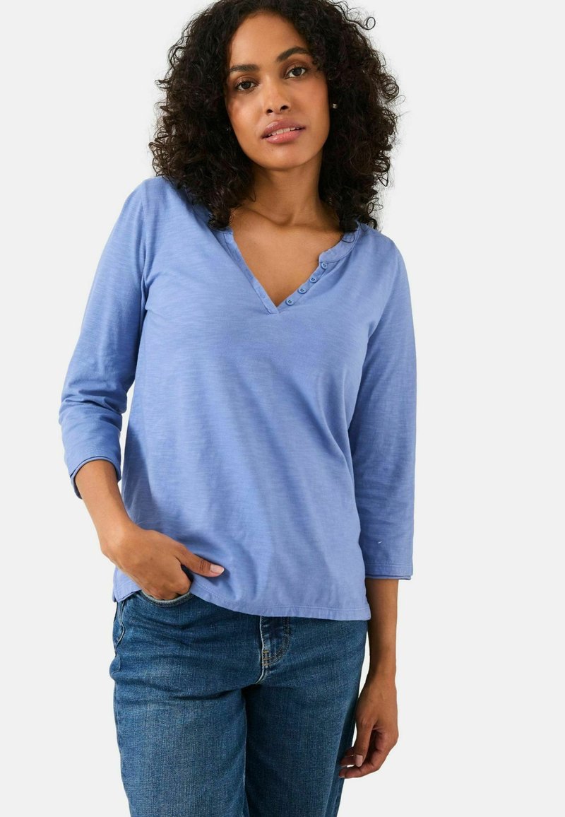 Woman with curly hair wearing a light blue 3/4 sleeve henley shirt and blue jeans, standing with one hand in her pocket.
