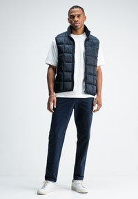 Schwarze Steppweste mit gesteppter Optik, getragen über einem weißen T-Shirt, kombiniert mit marineblauen Hosen und weißen Sneakers. Einfaches und lässiges Design.