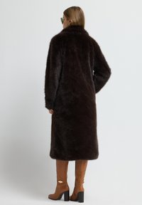 Abrigo de pelaje sintético marrón con cuello alto y mangas largas, que presenta una textura suave. Combinado con botas de tacón marrón hasta la rodilla y gafas de sol verdes.