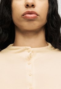 Κοντινό πλάνο του κάτω μέρους του προσώπου και του λαιμού μιας γυναίκας, που δείχνει ένα μπεζ πουκάμισο με κουμπιά και ένα χρυσό κρίκο στη ρινική της δεξιά πλευρά.