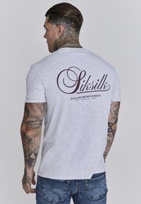 Pilkas trumpas rankovės marškinėliai su dideliu bordo logotipu ant nugaros. Pagaminta iš medvilnės, su glotnia tekstūra. Suderinta su mėlynomis džinsomis.