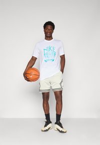 Hvid Nike t-shirt med turkist logo, hvide perforerede shorts med sorte kanter, sorte sokker og beige basketballsko, holdende en basketball.